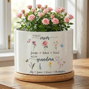 Puede incluir: Una maceta de cerámica blanca con las palabras "First Mom Now Grandma" y nombres. La maceta contiene rosas rosas y se asienta sobre una base de madera. Mariposas e ilustraciones florales decoran la maceta.