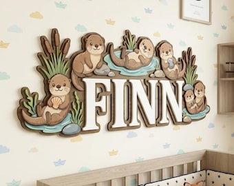 May include: A wooden wall decoration featuring the name "FINN" in white letters, with a brown border. The design includes cartoon otters, water, and reeds. The background is a light-colored wall with a pattern of clouds and paper boats.