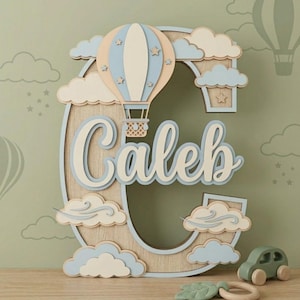 Puede incluir: Una letra C de madera grande con un diseño en capas que incluye un globo aerostático, nubes y el nombre Caleb en letra cursiva. La pieza está decorada en tonos azul claro, crema y madera natural con detalles de estrellas.