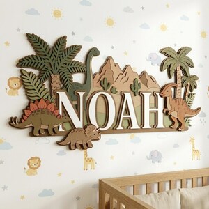 Pu&ograve; includere: Decorazione da parete in legno con il nome "NOAH" a tema dinosauri. Le lettere sono bianche con un bordo marrone chiaro. Il design include vari dinosauri, palme, montagne e cactus nei toni del verde, marrone e arancione.