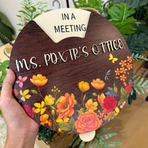 Op de afbeelding: Een ronde houten kantoorbord met een donkerbruine achtergrond en bloemenrand. Het bord leest "IN A MEETING" en "MS. POXTR'S OFFICE" in witte letters. De bloemenrand heeft oranje, gele en rode bloemen.