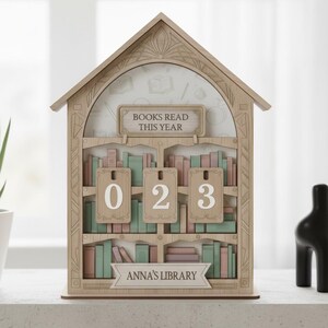 Könnte beinhalten: Holzregal-förmiger Kalender mit dem Text "BOOKS READ THIS YEAR" und "ANNA'S LIBRARY". Der Kalender zeigt die Zahlen "023" auf hängenden Etiketten, mit Bücherregalen in Pastellfarben.