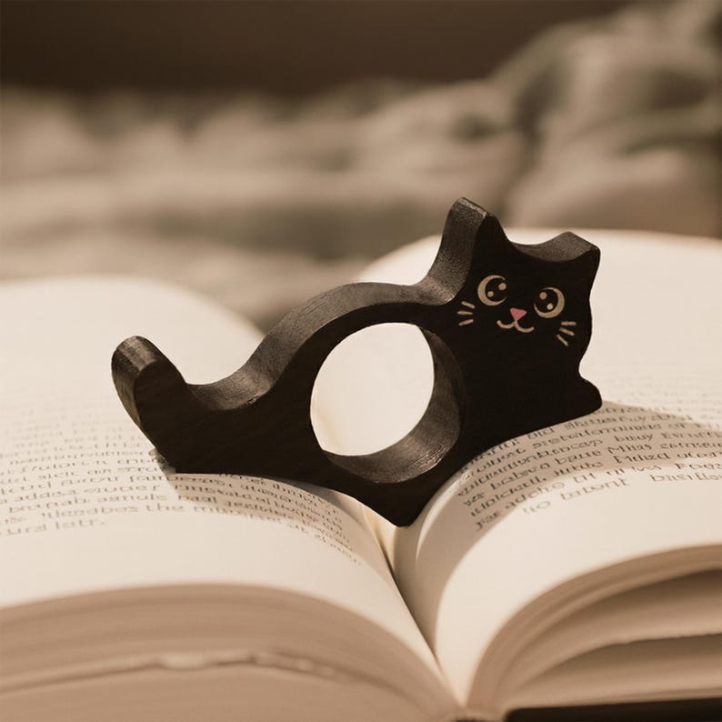 Puede incluir: Un marcap&aacute;ginas de madera negra con forma de gato descansa sobre las p&aacute;ginas abiertas de un libro. El marcap&aacute;ginas tiene una abertura circular y una cara de gato sonriente con bigotes blancos y boca rosa. Las p&aacute;ginas del libro est&aacute;n llenas de texto peque&ntilde;o.