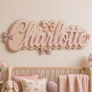 Puede incluir: Un letrero de pared de madera en capas con el nombre Charlotte en letra cursiva. Las letras son de color rosa claro con un borde de madera natural. El letrero está decorado con lazos rosas tridimensionales y pequeños detalles perlados.