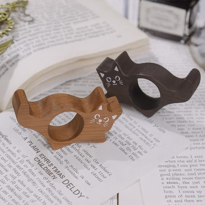 Puede incluir: Dos servilleteros de madera con forma de gato. Uno es marr&oacute;n claro, el otro es marr&oacute;n oscuro. Cada uno tiene una cara de gato pintada de blanco. Est&aacute;n sobre un libro con texto impreso.