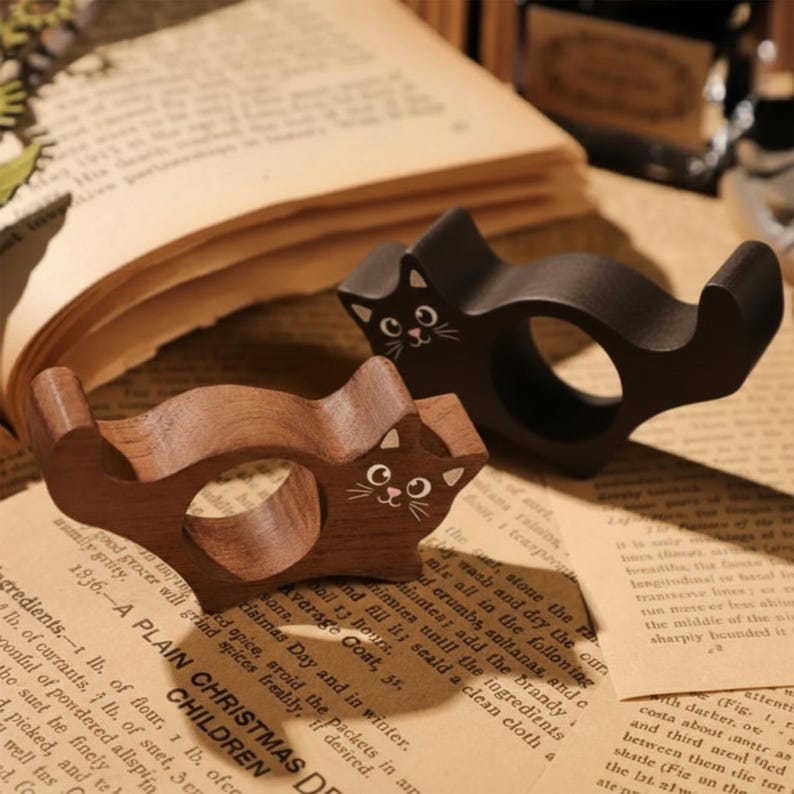 Puede incluir: Dos servilleteros de madera con forma de gato. Uno es marr&oacute;n oscuro, el otro negro. Ambos tienen caras de gato pintadas de blanco. Los servilleteros est&aacute;n sobre un libro abierto con p&aacute;ginas vintage.