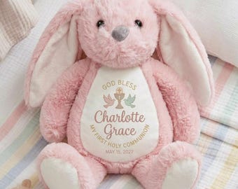 Puede incluir: Un conejo de peluche rosa con orejas largas y una nariz gris. El conejo tiene un parche blanco en el pecho con el texto "GOD BLESS", dos palomas, un cáliz, el nombre "Charlotte Grace" y "MY FIRST HOLY COMMUNION MAY 15, 2027".