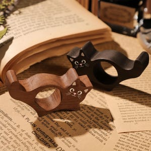 Puede incluir: Dos servilleteros de madera con forma de gato. Uno es marr&oacute;n oscuro, el otro negro. Ambos tienen rasgos faciales pintados de blanco. Est&aacute;n sobre una p&aacute;gina de libro vintage. Estos anillos decorativos a&ntilde;aden un toque caprichoso a cualquier mesa.