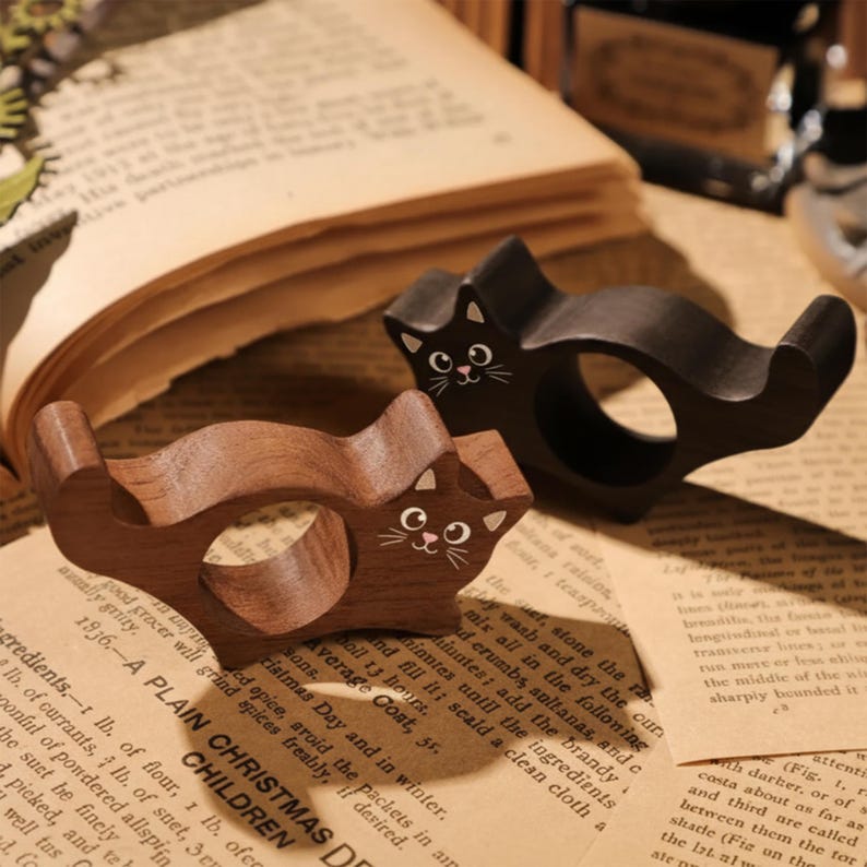 Puede incluir: Dos servilleteros de madera con forma de gato. Uno es marr&oacute;n oscuro, el otro negro. Ambos tienen rasgos faciales pintados de blanco. Est&aacute;n sobre una p&aacute;gina de libro vintage. Estos anillos decorativos a&ntilde;aden un toque caprichoso a cualquier mesa.
