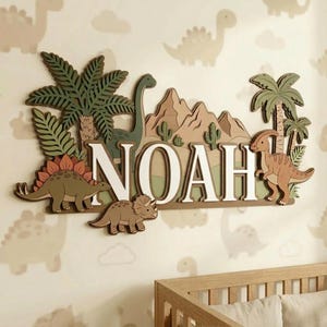 Pu&ograve; includere: Decorazione murale in legno con il nome "NOAH" in lettere bianche, circondato da un tema di dinosauri. Il design include vari dinosauri, palme, montagne e cactus in tonalit&agrave; di verde, marrone e arancione. Lo sfondo &egrave; beige chiaro con silhouette di dinosauri.