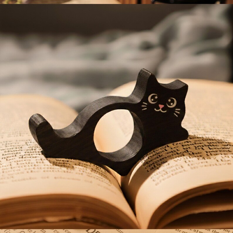 Puede incluir: Un marcap&aacute;ginas de madera negra con forma de gato y una cara linda, colocado sobre las p&aacute;ginas abiertas de un libro. El marcap&aacute;ginas tiene una abertura circular y est&aacute; dise&ntilde;ado para colocarse en el borde del libro. Las p&aacute;ginas del libro est&aacute;n envejecidas y amarillentas.