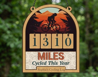 May include: Wooden cycling log sign with a silhouette of a cyclist riding a bike against a sunset backdrop. The sign displays "1310 MILES CYCLED THIS YEAR" and "Sophia's Cycling Log". The sign is hanging from a chain.