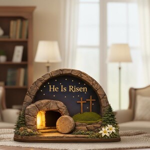 May include: A decorative Easter scene featuring a stone-like tomb with the words "He Is Risen" on a dark blue background. The scene includes two crosses, a stone, and a lit interior, with small trees and flowers.