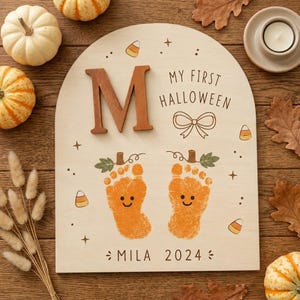 以下が含まれることがあります： 大きな茶色の文字M、"MY FIRST HALLOWEEN"のテキスト、リボンの線画、笑顔が描かれた2つのオレンジ色の足跡のイラストが特徴のアーチ型木製看板。下部には"MILA 2024"のテキストとキャンディコーンのアイコンが含まれています。