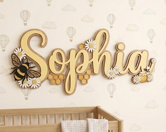 Puede incluir: Un letrero decorativo de madera para pared con el nombre Sophia en letras cursivas grandes de color amarillo claro. El diseño incluye un abejorro negro y amarillo, flores de margarita blancas y patrones de panal amarillos integrados.