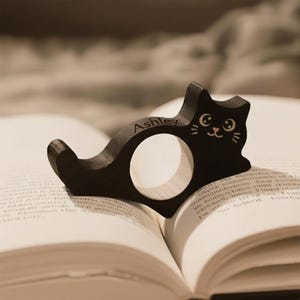 Puede incluir: Un sujetap&aacute;ginas de libro con forma de gato de madera negra con el nombre "Ashley" grabado. El gato tiene una cara sonriente y descansa sobre las p&aacute;ginas de un libro abierto. El soporte est&aacute; dise&ntilde;ado para mantener el libro abierto.