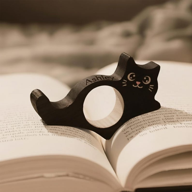 Puede incluir: Un sujetap&aacute;ginas de libro con forma de gato de madera negra con el nombre "Ashley" grabado. El gato tiene una cara sonriente y descansa sobre las p&aacute;ginas de un libro abierto. El soporte est&aacute; dise&ntilde;ado para mantener el libro abierto.