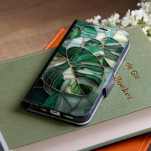 May include: A black phone case with a green and white stained glass design featuring a monstera leaf pattern. The case is made of leather and has a magnetic closure. The phone case is resting on a green book.