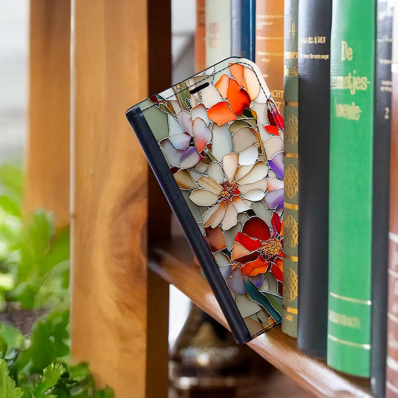 May include: A floral-patterned phone case with a black border, featuring a stained-glass design of red, orange, and white flowers. The case is propped up against a bookshelf with various books.
