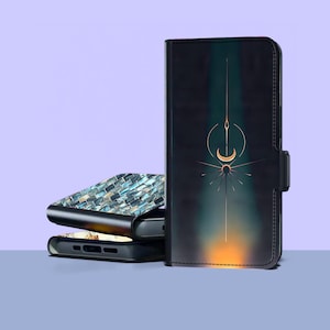 Op de afbeelding: Zwarte telefoonhoes met een hemels ontwerp met een gouden halve maan en zonnestralen. De hoes heeft een magnetische sluiting. Twee extra telefoonhoesjes zijn erachter gestapeld, een met een mozaïekpatroon.