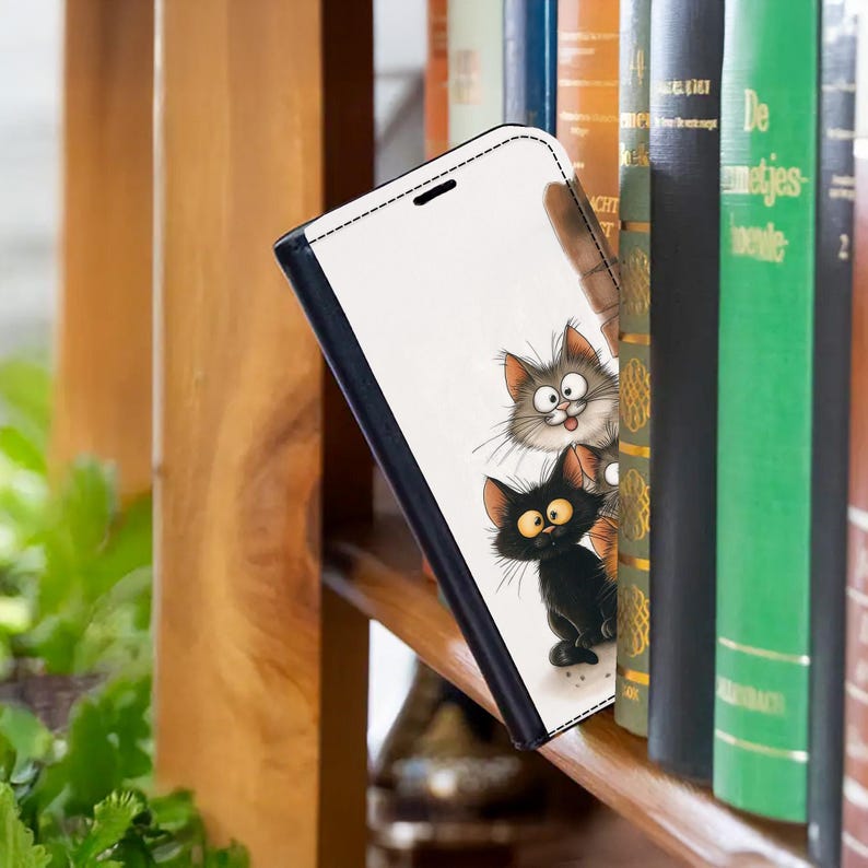 Pode incluir: Uma capa de telefone branca com uma borda preta com uma ilustra&ccedil;&atilde;o de desenho animado de dois gatos. Um gato &eacute; cinza e o outro &eacute; preto. A capa est&aacute; encostada em uma estante.