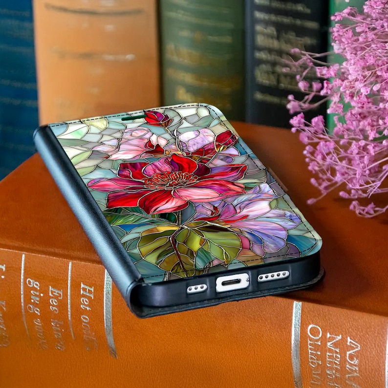 Peut inclure: Un &eacute;tui de t&eacute;l&eacute;phone noir avec un motif floral en vitrail repr&eacute;sentant des fleurs rouges, roses et violettes. L'&eacute;tui est ouvert, r&eacute;v&eacute;lant les ports et les boutons du t&eacute;l&eacute;phone. Le motif est sur fond turquoise.