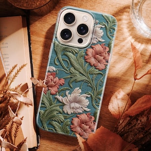 Puede incluir: Una funda de teléfono turquesa con un diseño floral con flores rosas y blancas y hojas verdes. La funda tiene un borde transparente y un recorte para la cámara. La funda está sobre una superficie de madera.
