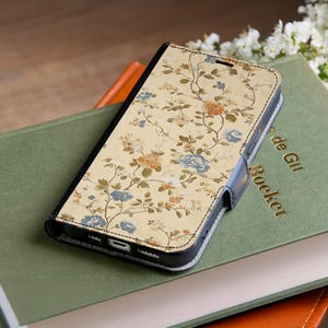 Könnte beinhalten: Eine Handyhülle mit floralem Muster, cremefarbenem Hintergrund und einem Muster aus blauen, orangefarbenen und gelben Blumen. Die Hülle ist geschlossen und hat einen schwarzen Rand und einen Magnetverschluss. Die Hülle liegt auf einem grünen Buch.