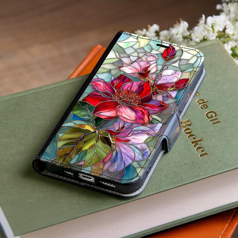 Peut inclure: Un &eacute;tui de t&eacute;l&eacute;phone noir avec un motif floral en vitrail vibrant. L'&oelig;uvre d'art pr&eacute;sente une grande fleur rouge avec des fleurs roses et violettes, des feuilles vertes et un fond bleu clair. L'&eacute;tui est ouvert, r&eacute;v&eacute;lant l'&eacute;cran du t&eacute;l&eacute;phone.