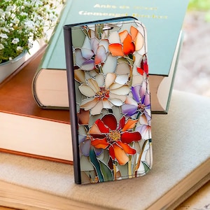 May include: A floral phone case featuring a stained-glass design with white, orange, and purple flowers. The case is black with a magnetic closure. The case is resting on a stack of books.