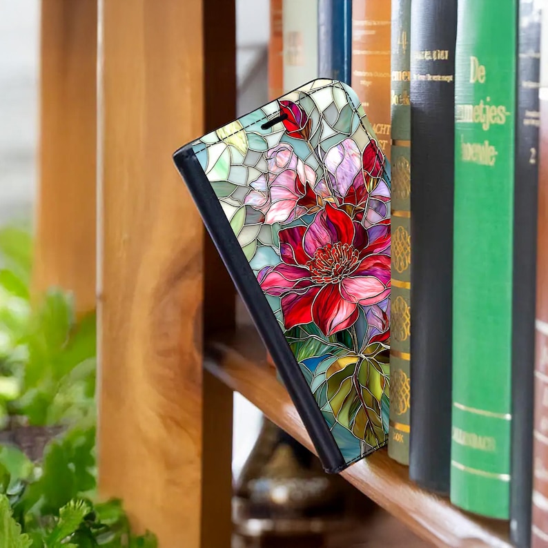 Peut inclure: Un &eacute;tui de t&eacute;l&eacute;phone noir avec un motif floral en vitrail aux teintes rouge, rose et verte. L'&eacute;tui est ouvert, r&eacute;v&eacute;lant l'int&eacute;rieur. Le motif comprend une grande fleur rouge.