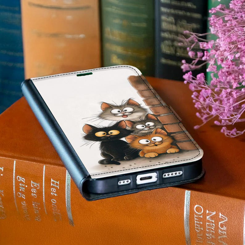 Pode incluir: Capa de telefone preta com uma ilustra&ccedil;&atilde;o de desenho animado de quatro gatos em v&aacute;rias cores. O design inclui uma parede de tijolos e um fundo branco. A capa est&aacute; aberta, revelando a tela do telefone.
