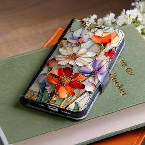 May include: A black phone case with a colorful floral design. The case features a stained-glass style pattern with flowers in shades of red, orange, purple, and white. The case is resting on a green book.