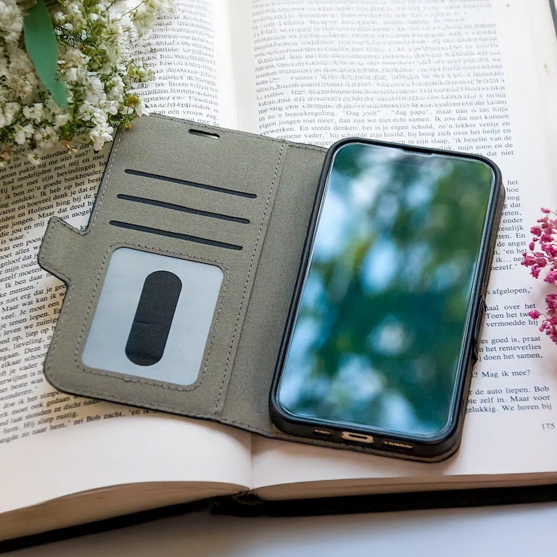 Pode incluir: Uma capa de telefone verde-oliva aberta com ranhuras para cart&otilde;es e uma janela de identifica&ccedil;&atilde;o transparente, segurando um smartphone preto. A capa est&aacute; em um livro aberto com texto, com flores ao fundo.