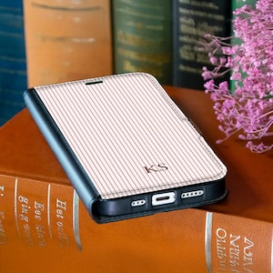 Peut inclure: Une coque de téléphone noire avec un rabat à fines rayures rose clair et blanches, portant les lettres KS gravées au centre. L'étui présente une découpe pour le port de charge et des orifices pour haut-parleurs sur le bord inférieur.