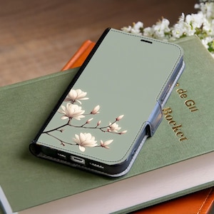 May include: A phone case with a floral design. The case is light green with a black border and a white floral branch. The case is open, revealing the phone screen. The case is resting on a green book.