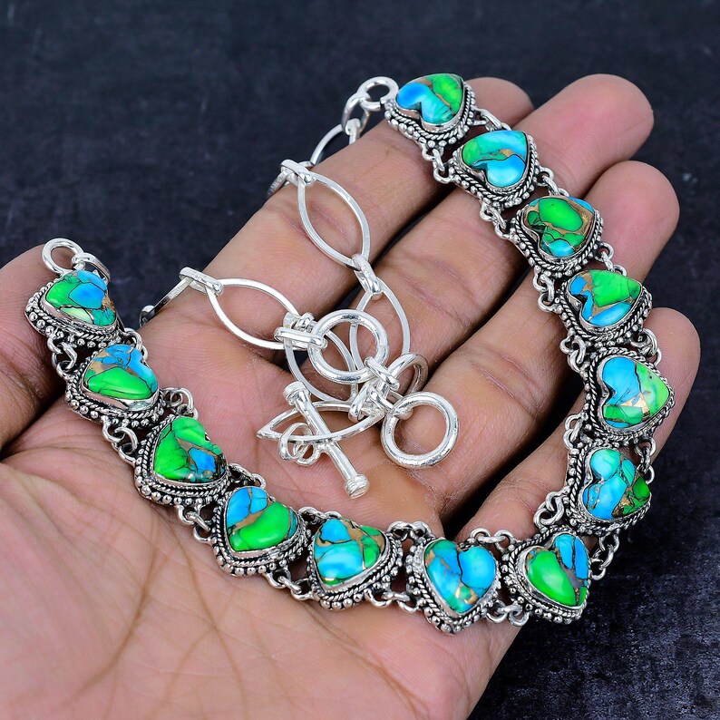 May include: A silver-toned bracelet featuring heart-shaped turquoise and green stones. The stones are set in a decorative, ornate silver setting. The bracelet has a toggle clasp and is displayed on a hand.