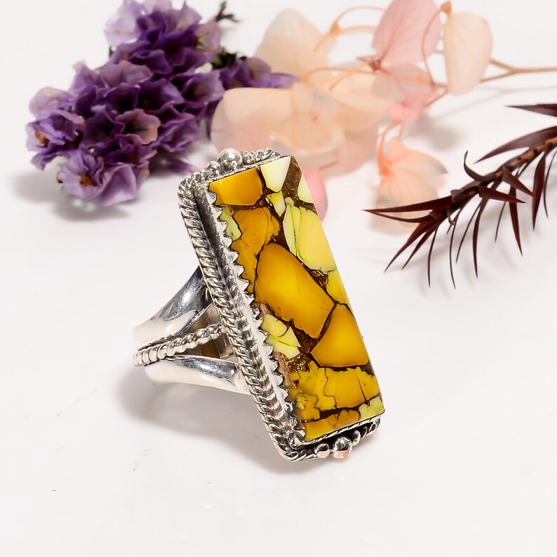 May include: A silver ring with a large, rectangular stone with a yellow and brown pattern. The stone is set in a decorative silver frame with a braided edge. The ring is displayed with purple and pink flowers.