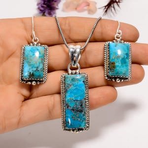 May include: A jewelry set featuring a pendant necklace and matching earrings. Each piece has a rectangular turquoise stone set in a silver-toned frame with a rope-like border. The turquoise has blue and brown marbling.
