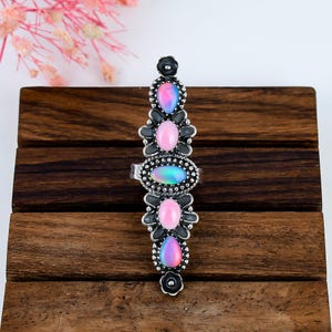 Peut inclure: Une longue bague argentée présentant une disposition verticale de cinq pierres cabochons. Les pierres alternent entre des formes de goutte iridescentes et des formes ovales roses unies, serties dans une monture décorative avec des fleurs.