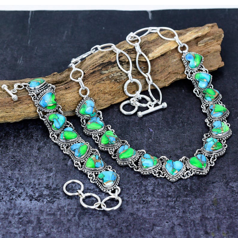 May include: A silver necklace and bracelet set featuring heart-shaped turquoise and green stones. The stones are set in a decorative silver frame and linked with a silver chain. The necklace has a toggle clasp.