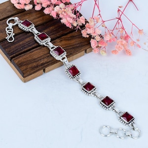 May include: A silver bracelet featuring square, deep red gemstones. The bracelet has an ornate design with small, beaded accents around each stone. The clasp is a simple hook and eye closure. The jewelry is displayed on a wooden surface.