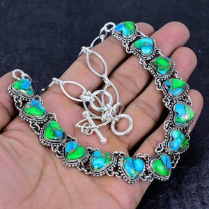 May include: A silver-toned bracelet featuring heart-shaped turquoise and green stones. The stones are set in a decorative, ornate silver setting. The bracelet has a toggle clasp and is displayed on a hand.