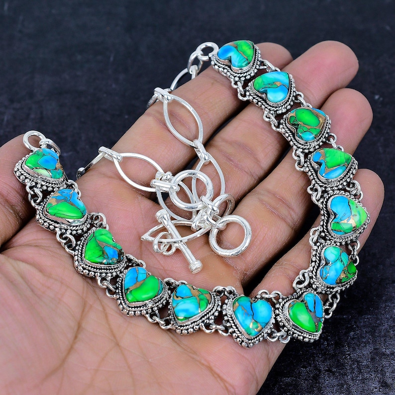 May include: A silver-toned bracelet featuring heart-shaped turquoise and green stones. The stones are set in a decorative, ornate silver setting. The bracelet has a toggle clasp and is displayed on a hand.