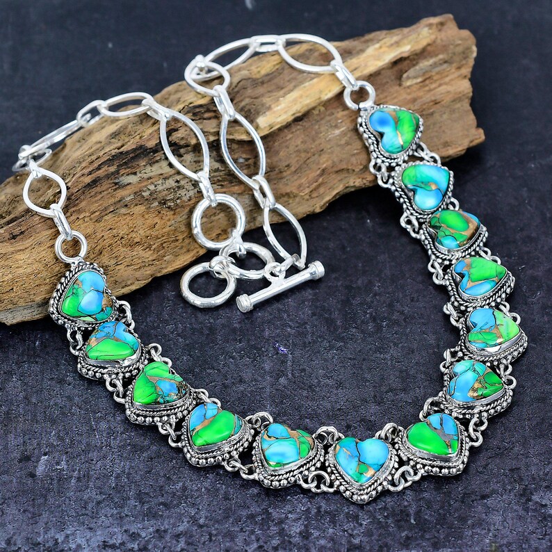 May include: A silver-toned necklace featuring heart-shaped turquoise and green stones. The stones are set in ornate, detailed bezels and linked together with a chain. The necklace has a toggle clasp and is displayed on a wooden surface.