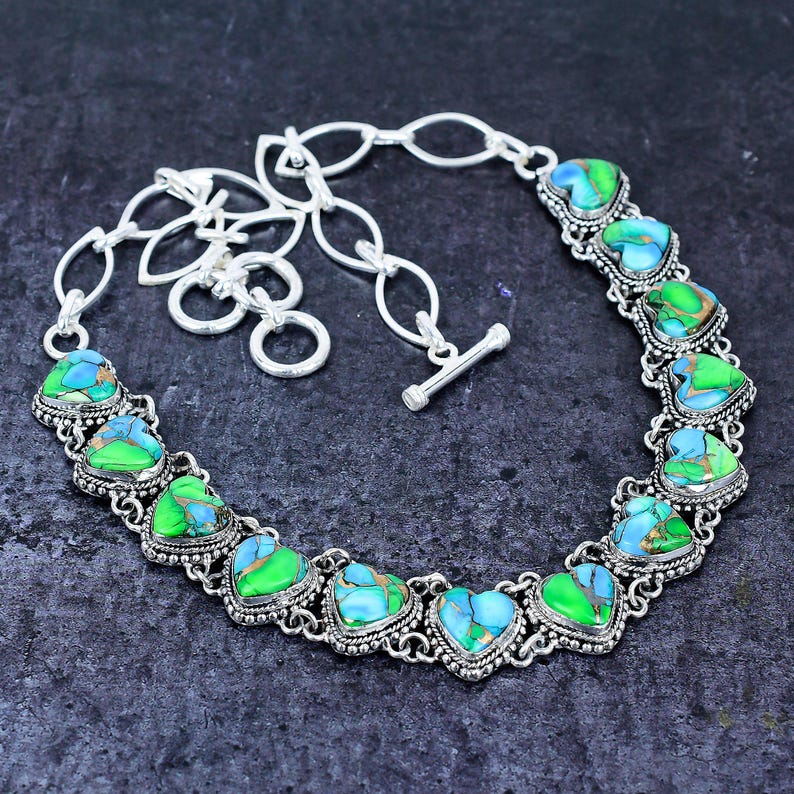 May include: A silver-toned necklace featuring heart-shaped turquoise and green stones. The stones are set in a decorative silver frame and connected by a chain. The necklace has a toggle clasp.
