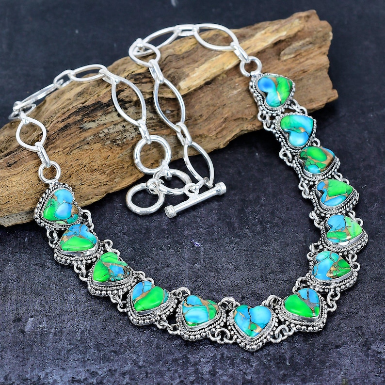 May include: A silver-toned necklace featuring heart-shaped turquoise and green stones. The stones are set in ornate, detailed bezels and linked together with a chain. The necklace has a toggle clasp and is displayed on a wooden surface.