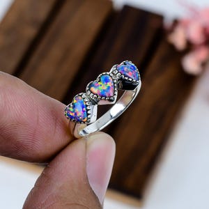 May include: A silver ring featuring three heart-shaped opal stones. The opals display a vibrant array of colors, including blue, green, and yellow. The ring's band is a polished silver, and the hearts are set in a decorative bezel.