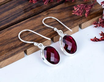 May include: Silver drop earrings featuring oval, faceted, deep red gemstones. The earrings are presented on a wooden surface, with dried red foliage in the background. The gemstones have a polished, reflective surface.
