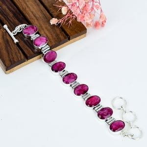 Könnte beinhalten: Ein Silberarmband mit neun ovalen, facettierten magentafarbenen Edelsteinen, die miteinander verbunden sind. Das Armband verfügt über einen Knebelverschluss an einem Ende und drei kreisförmige Ringe am anderen Ende zur Größenanpassung.